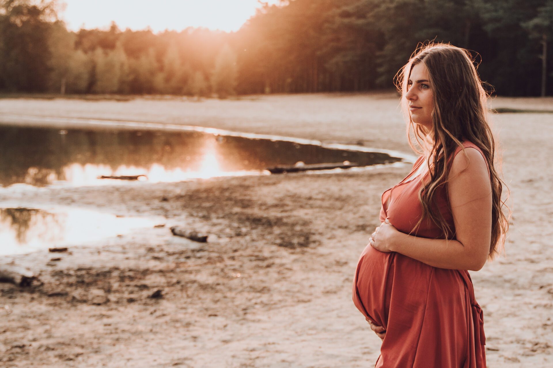 Zwangerschapsfotoshoot door flow photography, zwangere kijkend naar ondergaande zon naast een meer